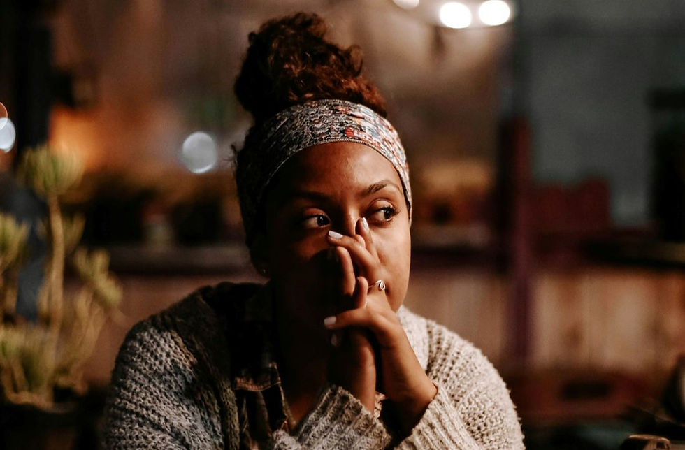 Woman with contemplative expression, wearing a patterned headband and knit sweater, resting her hands on her face in a dimly lit room.