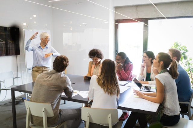Older man presents to a diverse team around a conference table in a bright modern meeting room, all listening intently.