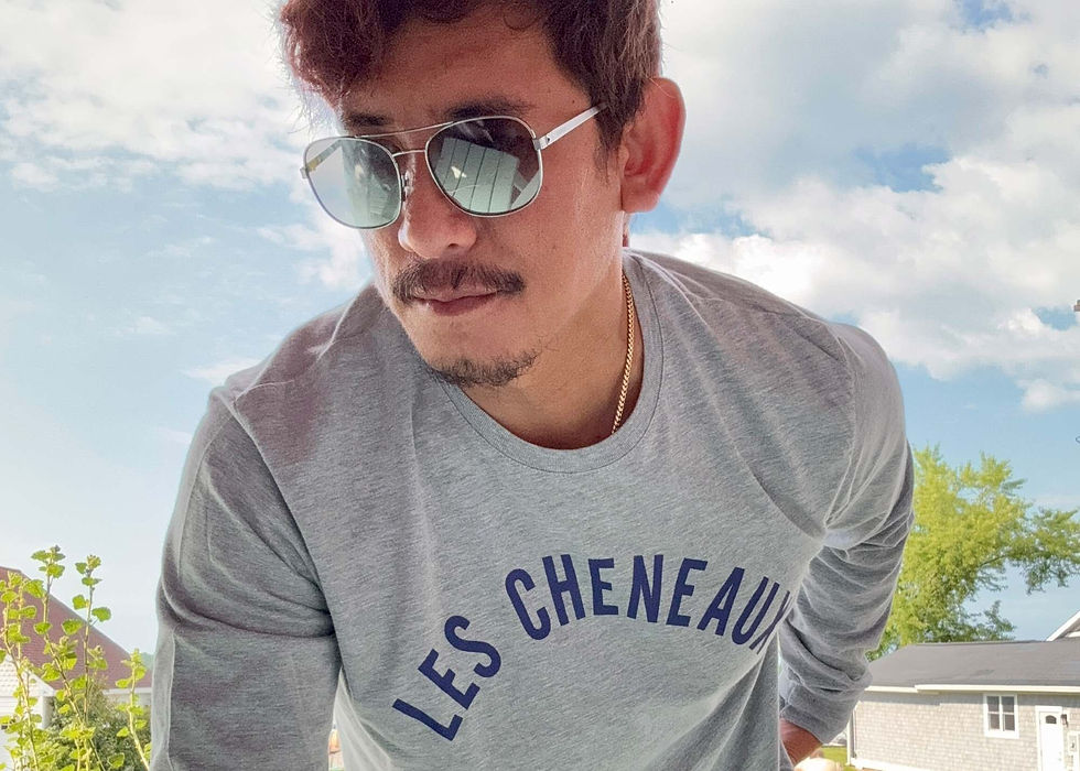 Man wearing sunglasses and a gray "LES CHENEAUX" shirt leans forward, with a clear sky and houses in the background. Casual vibe.