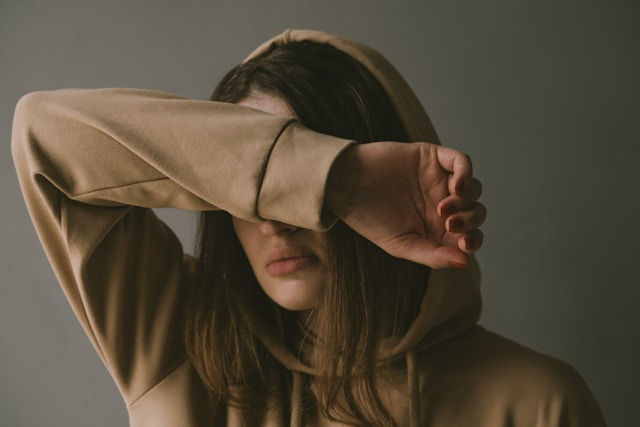 A close-up shot of a woman wearing a beige hoodie, her arm covering her eyes. The lighting is subdued, and the overall mood is contemplative or melancholic.