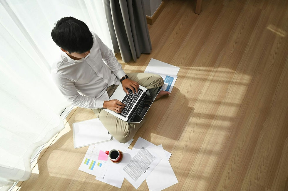 Person sitting on wooden floor, using laptop, surrounded by papers and a red mug. Bright curtains in the background, creating a calm atmosphere.