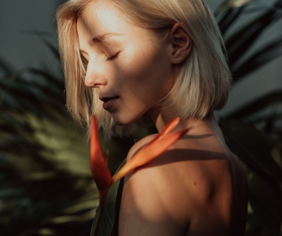 A woman with closed eyes stands in soft sunlight, holding a tropical orange flower against her shoulder.