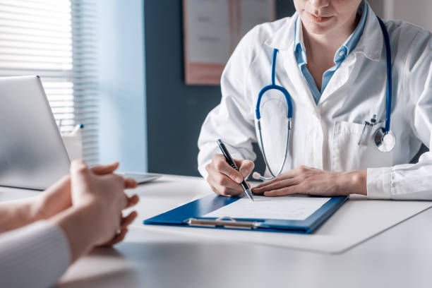 Doctor in white coat writing on clipboard, patient hands visible, office setting with laptop and documents. Bright, professional atmosphere.