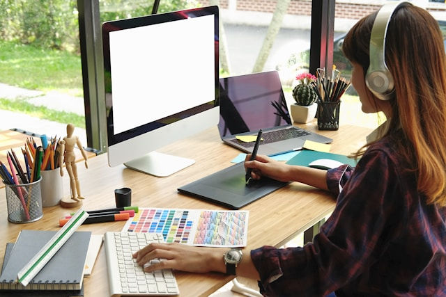 Woman wearing headphones draws on a tablet at a desk with an iMac, laptop, and art supplies by a bright window.