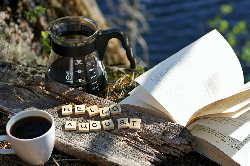 Coffee cup, glass carafe, and open book on a log by water, with letter tiles spelling HELLO AUGUST in a calm outdoor scene