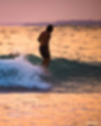 A person surfs a wave at sunset, surrounded by pink and orange hues. The ocean shimmers in the warm light. Signature reads "Sarah Lee."