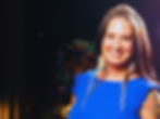 Smiling woman in a blue dress with braided hair and feather accessories, standing in a dimly lit room with a colorful lion painting behind her.