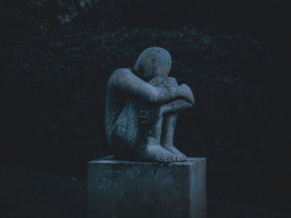 A stone sculpture of a seated figure hugging knees, on a pedestal. The setting is dark and moody, with a shadowy, leafy background.