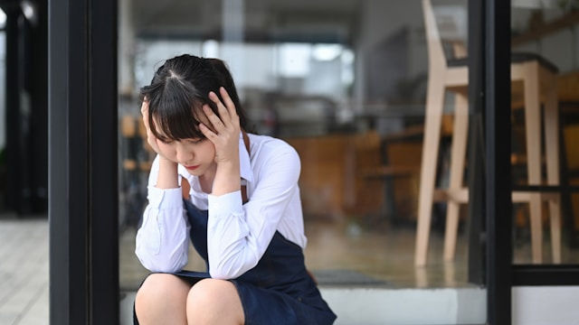 Young woman in a white shirt and dark apron sits outside a cafe, holding her head in her hands and looking worried.