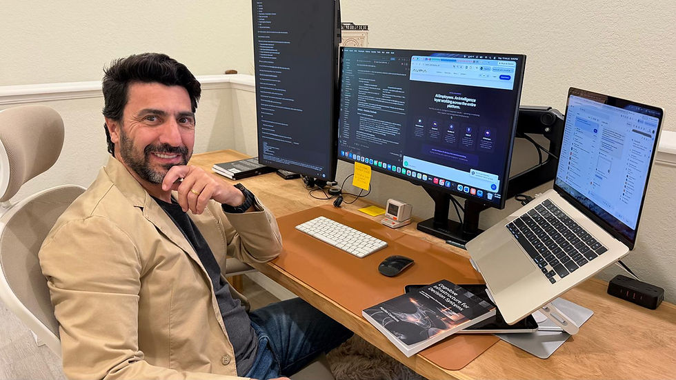 Smiling man at a desk with three monitors and a laptop showing apps and code; home office with books and keyboard.
