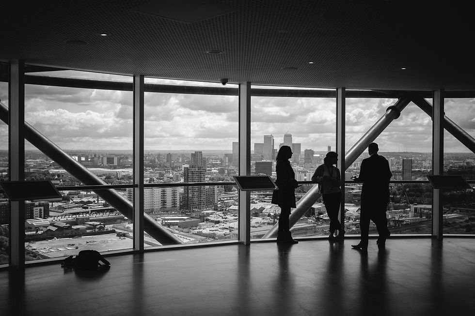 Silhouetted people stand and talk in front of large windows showing a cityscape. Overcast sky, modern interior, and an urban view.