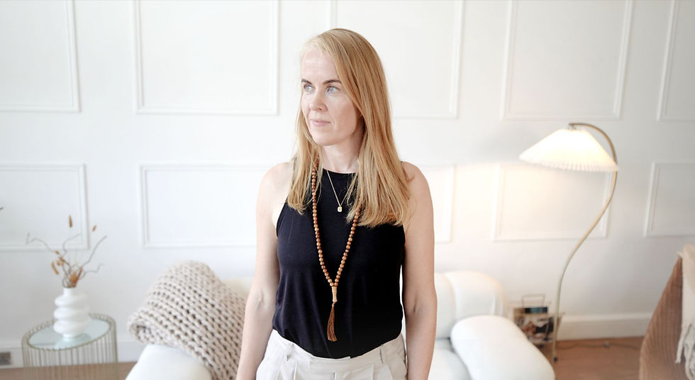 A woman in a black top stands in a bright room with white paneled walls, a floor lamp, and cozy decor. She appears calm and contemplative.