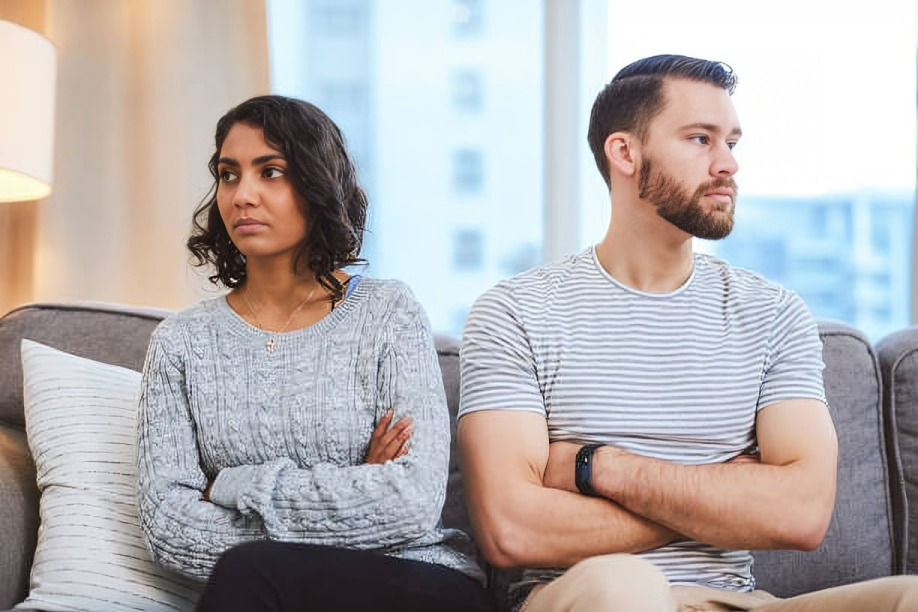 A couple sits on a couch with arms crossed, looking away from each other, indicating tension. Bright room with large window in background.