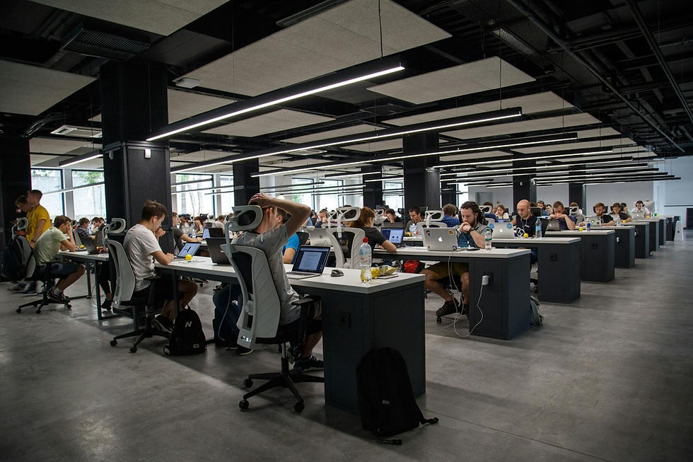 Open office with rows of people working at computers. Industrial ceiling, bright lights, modern desks. Appears busy and focused.