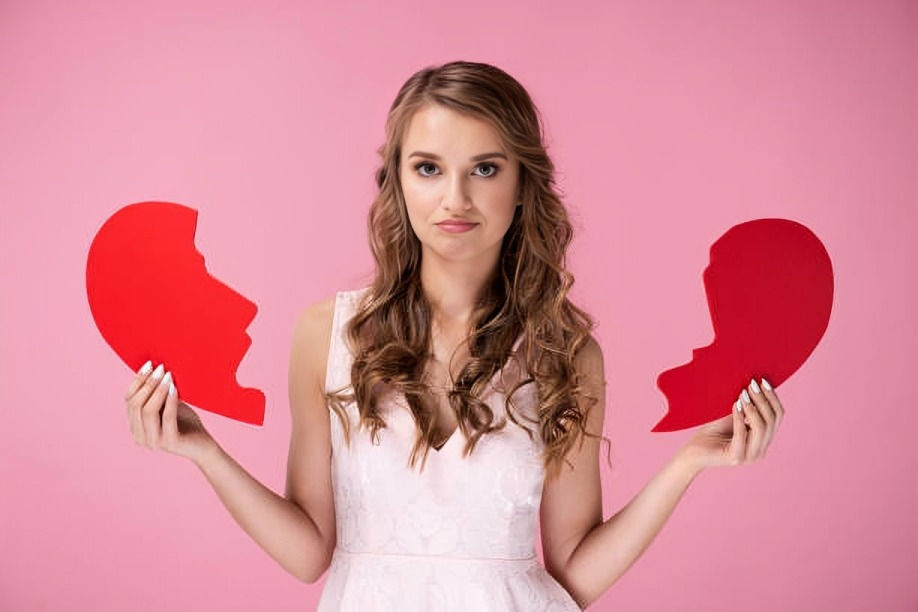 Woman in a white dress holds two red heart halves against a pink background, looking neutral or slightly sad.