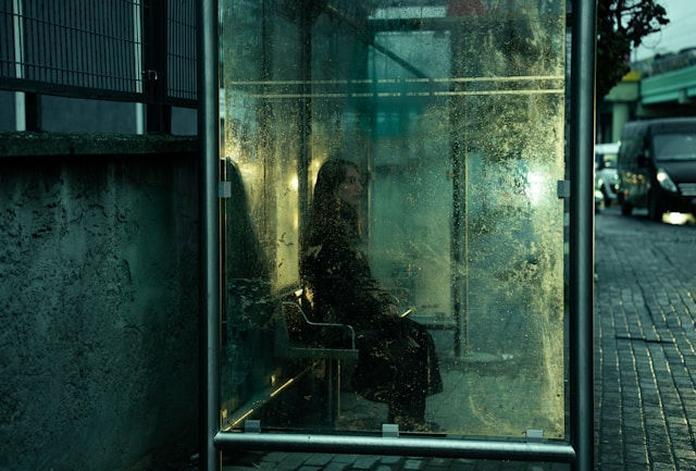 A woman sits inside a weathered glass bus stop, waiting.
