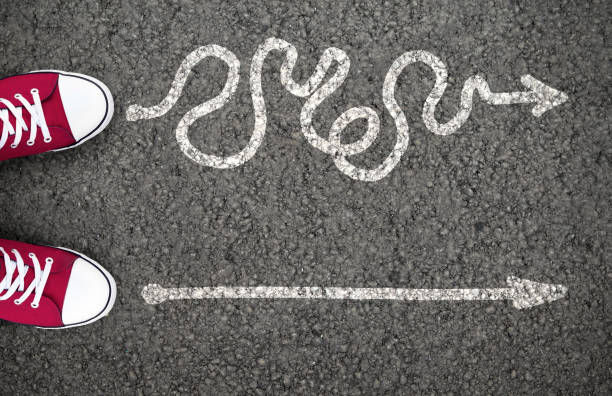 Red sneakers stand on asphalt facing two arrow paths: one straight, one winding. Choices depicted, mood of decision-making.