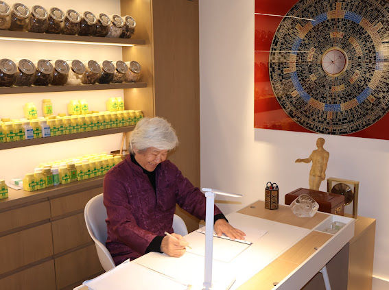 Elderly person in a purple jacket writing at a modern desk, surrounded by jars on shelves. A large circular artwork decorates the wall.