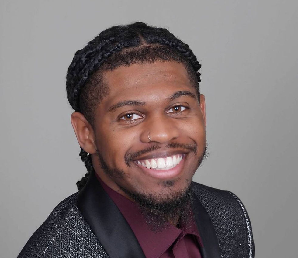 Smiling man with braided hair in a gray suit and maroon shirt against a plain gray background. He exudes a cheerful, friendly mood.