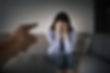 A woman sits on a gray sofa, covering her face with hands, expressing distress. A finger points at her from the foreground, suggesting conflict.