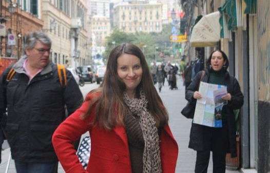 Photo of Nicoleen in a red coat posing confidently on a busy city street, with pedestrians walking in the background, including a man with a backpack and a person holding a map.