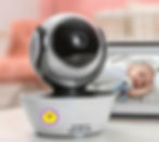 Baby monitor in foreground, black and white design. Screen shows a baby lying with stuffed toy. Soft pink and grey background.