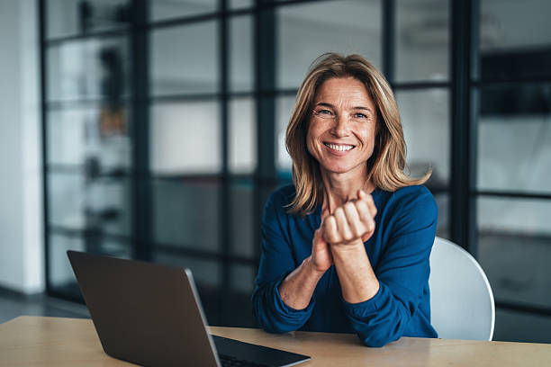 Smiling woman in a blue top sits at a laptop in a modern office, clasping her hands and looking cheerful.