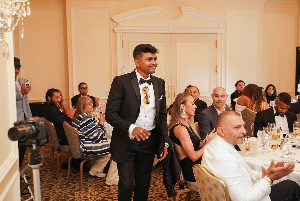 Man in a tuxedo stands smiling, receiving applause in an elegant banquet room. People sit at round tables, clapping. Mood is celebratory.