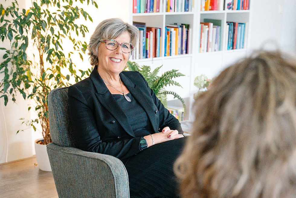 Woman in a black jacket smiles while seated in a chair. Bookshelves and plants are in the background, creating a cozy atmosphere.