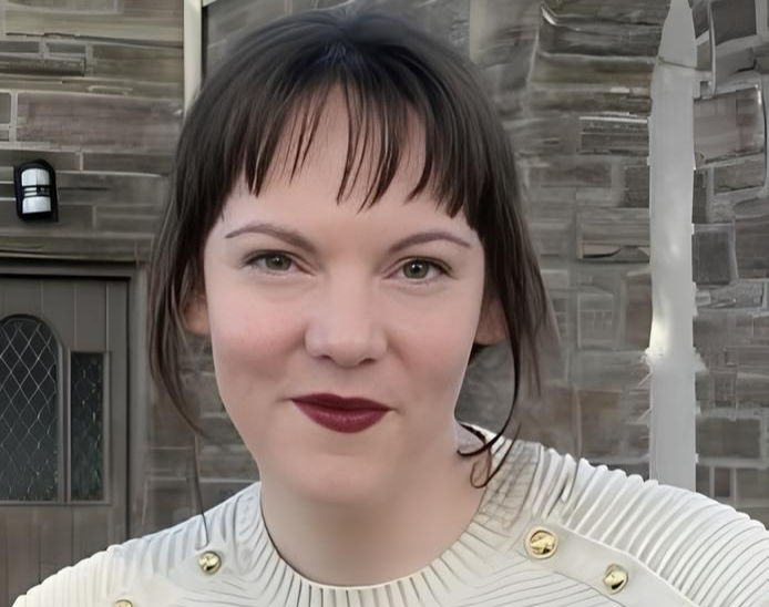 Isabella Presley with dark hair and red lipstick smiles in front of a stone building. She wears a white top with gold accents, creating a calm mood.
