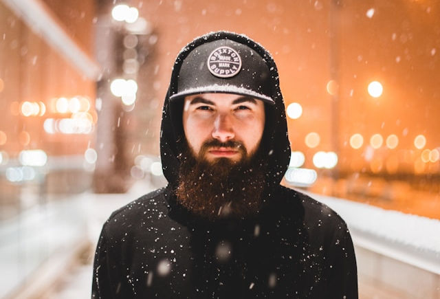 The photo features a bearded man wearing a black hoodie and a black cap, standing outdoors in a snowy urban setting at night.