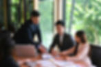 Business team in a bright meeting room reviews charts; one man points at papers beside a laptop, with green trees outside.