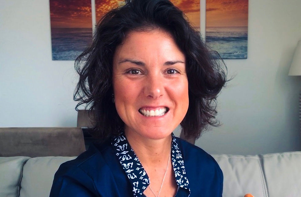Smiling woman in a navy shirt with a white pattern, sitting on a couch. Ocean sunset artwork on the wall in the background. Relaxed mood.
