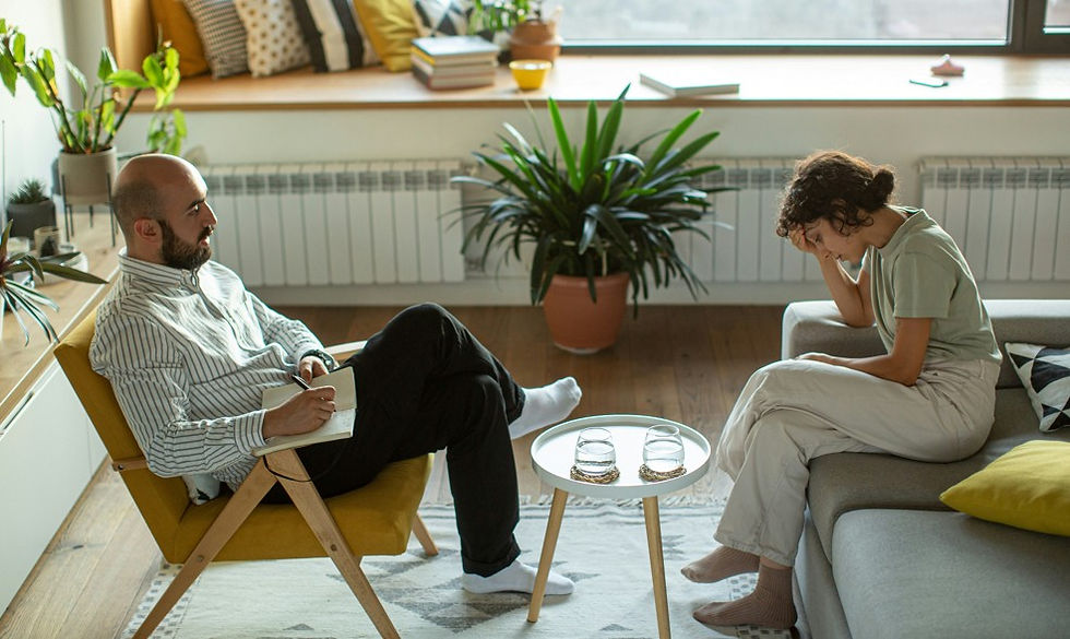 Therapist taking notes as a distressed woman sits on a sofa in a bright living room with plants and a small table.