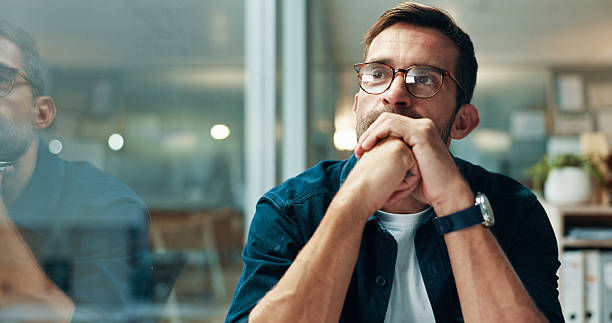 Thoughtful man with glasses sits in an office, hands clasped near his mouth, with soft reflections in the glass.