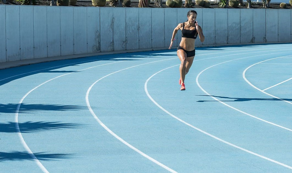 Woman sprinting on a blue track in a sports bra and shorts, with palm shadows and a concrete wall. Bright, sunny day.