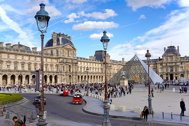 The photo shows the Louvre Museum in Paris with its iconic glass pyramid entrance, surrounded by historic architecture and a crowd of visitors under a bright blue sky.