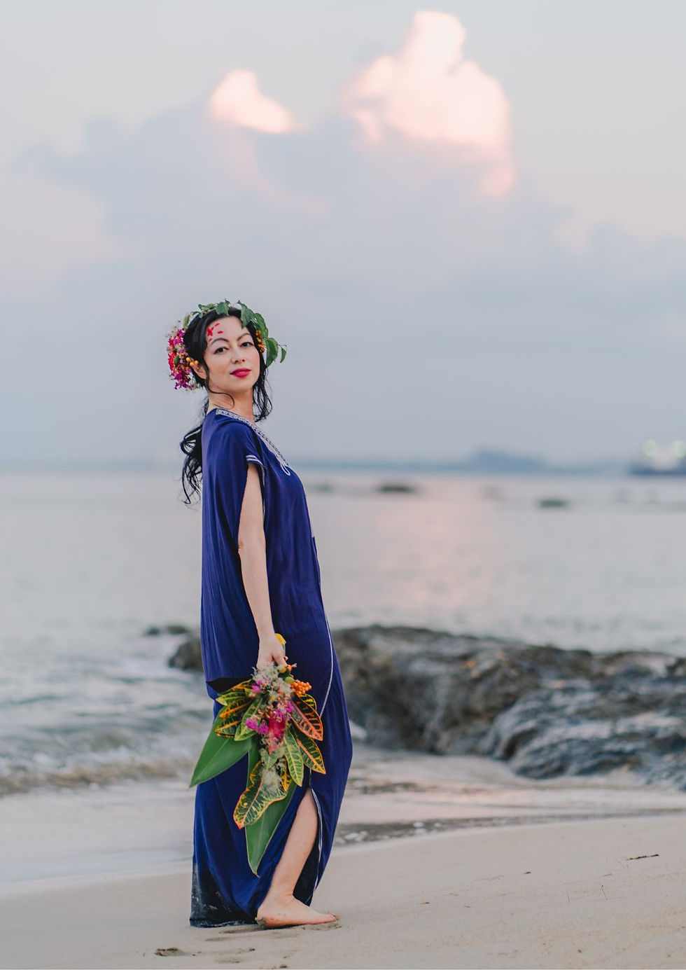 Woman in blue dress with floral crown holds bouquet on a beach. Soft pink clouds and a serene ocean create a tranquil setting.