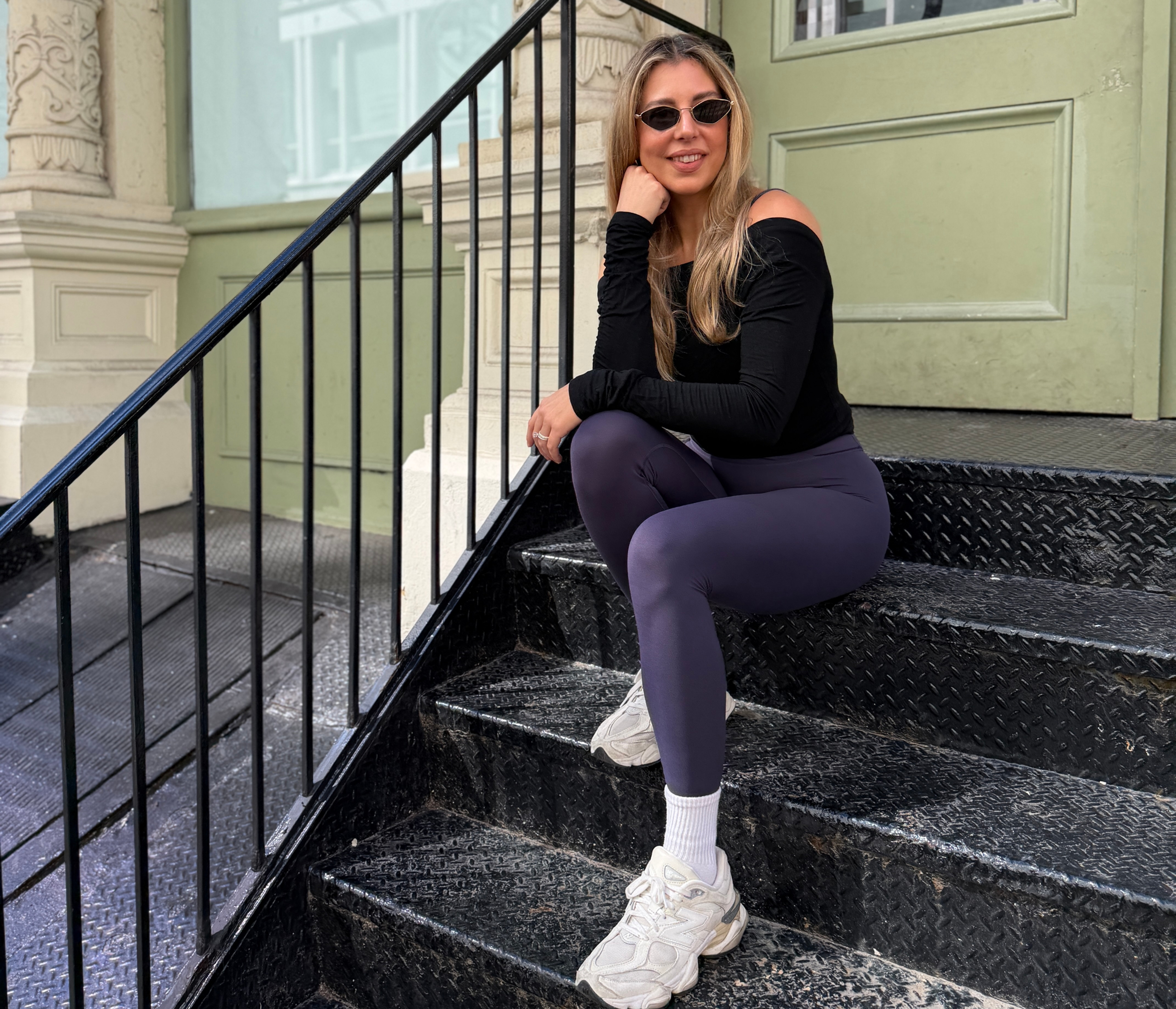 Woman in sunglasses sits on black steps outside a pale green building, smiling beside a door marked PUSH.