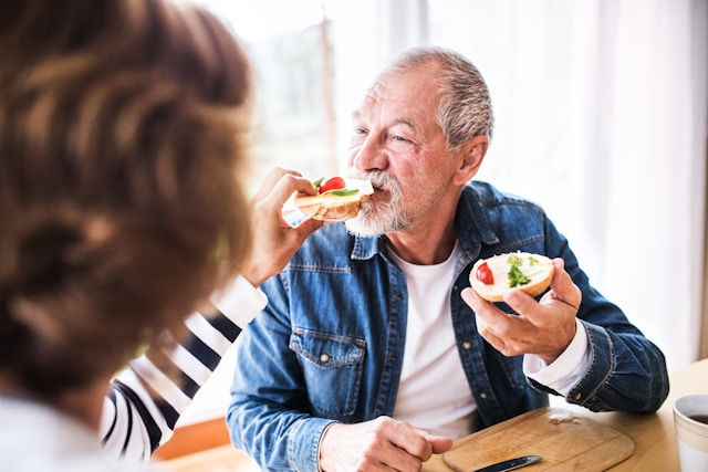 Man in denim jacket enjoying food, smiling. Someone in striped shirt offers him a bite. Bright indoor setting with natural light.