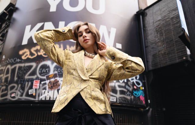 A confident woman in a gold patterned blazer poses in front of a graffiti-covered wall.