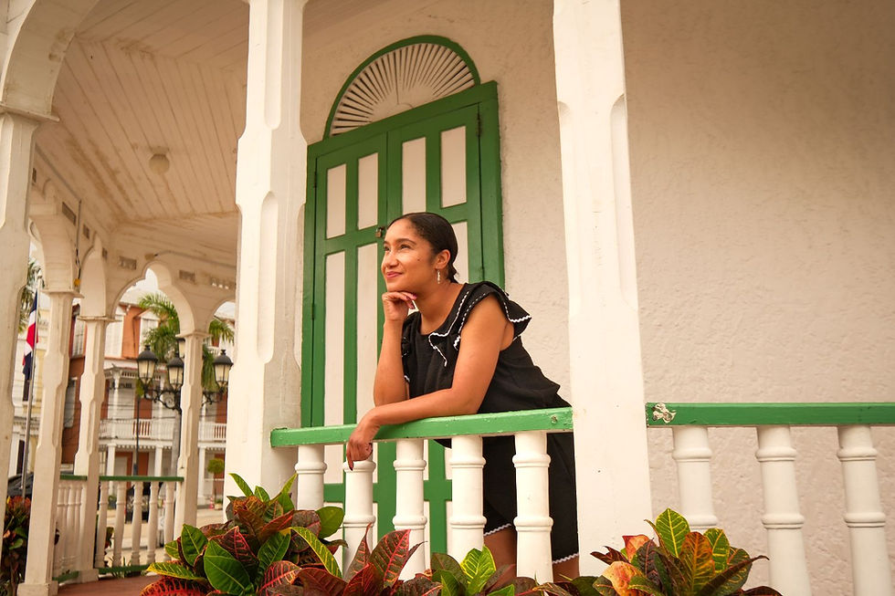 Woman in black dress leans on a white porch railing by a green door, smiling thoughtfully amid tropical plants.