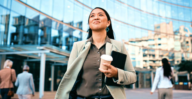 Businesswoman in a green blazer and black shirt holds a coffee and tablet, walking confidently outside a modern glass building, smiling.