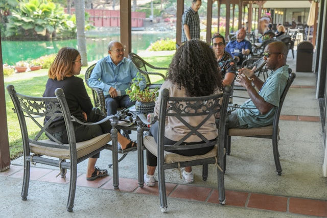 Five people sit in chairs around a table talking. The setting is a covered patio by a pond, with greenery and a relaxed atmosphere.