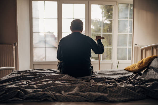 An elderly person sits on a bed with a cane, facing a large window. The room is dim with muted colors, creating a reflective mood.