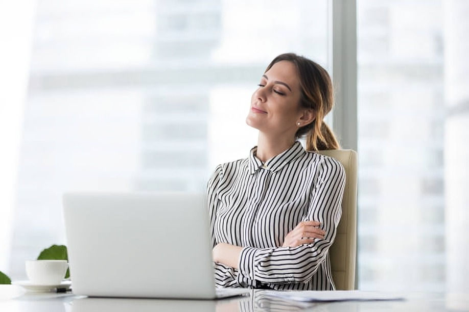 Woman in striped shirt sitting at a desk, eyes closed, smiling peacefully. Laptop and coffee cup on table. Bright office background.