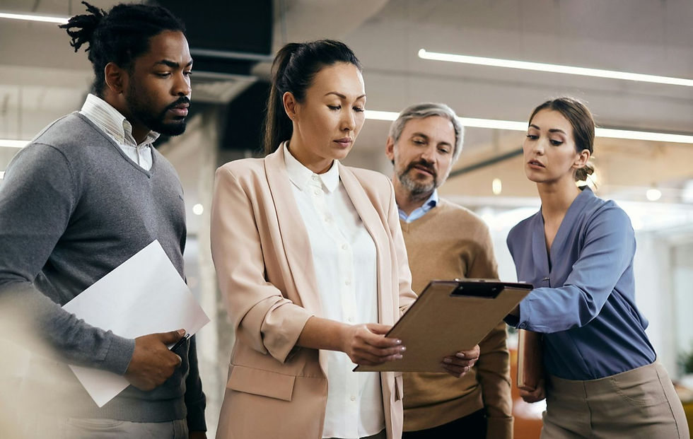 Four people in business attire discuss a clipboard in a modern office. They appear focused. Bright overhead lighting highlights the setting.