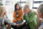 Elderly woman in green blazer, hands covering face, comforted by group. Bright room, supportive atmosphere.
