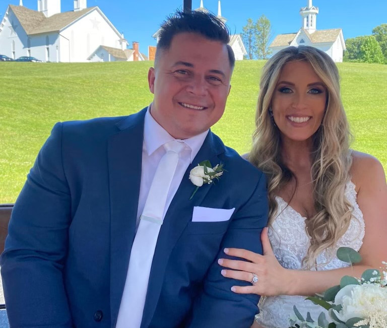 Smiling bride and groom in a golf cart at an outdoor wedding, with white church buildings on a sunny green hill behind them.