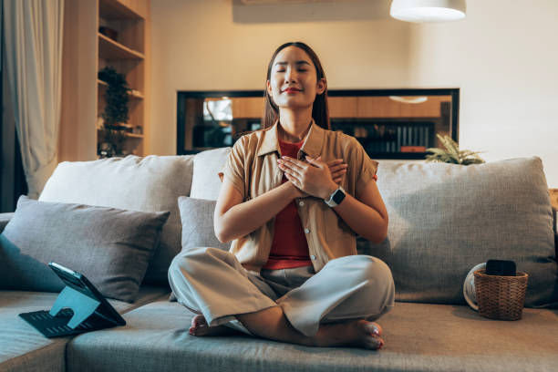 Woman meditating cross-legged on a sofa, eyes closed, hands on chest. Cozy living room with cushions, tablet, and warm lighting. Calm mood.